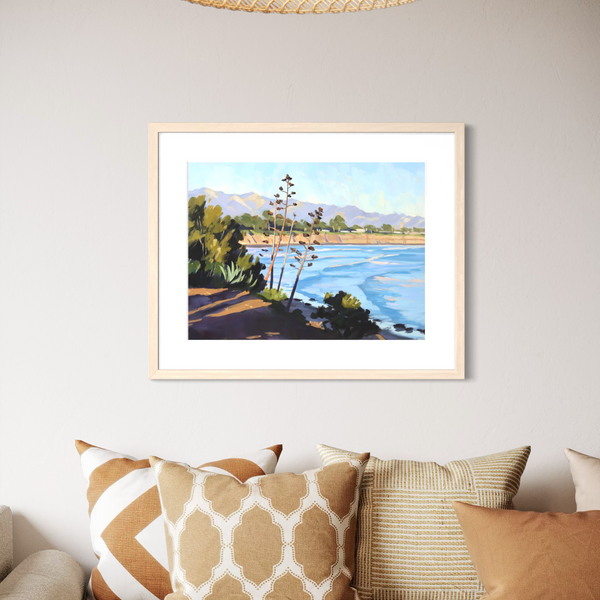 A framed landscape art print featuring a scenic river or lake with mountains, trees, and a shoreline, displayed on a light gray wall above a sofa with patterned cushions.