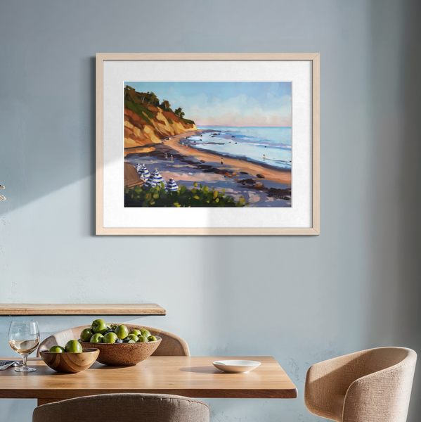 A framed coastal landscape painting hangs on a light blue-gray wall above a wooden dining table and upholstered chairs in a styled dining room.